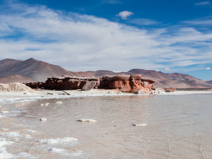 Red rocks beside the turquoise Aguas Calientes salt lake with volcanoes behind in northern Chile
